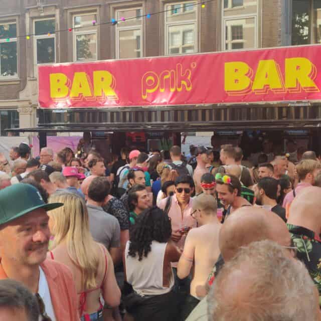 A crowded street party with many people in front of a large pink banner stating Prik Bar